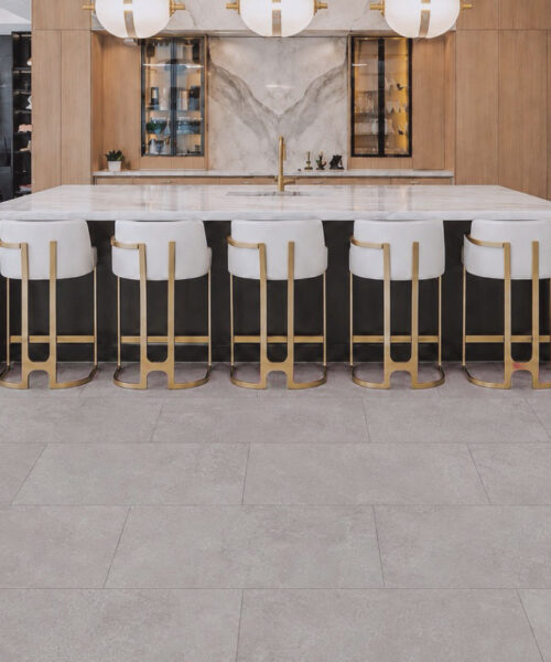 Bright Stone stone-look flooring covers a designer kitchen in large pale gray panels beneath a marble island and white barstools.