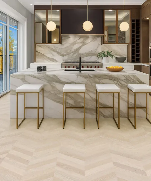 Clara Chevron - 7/16″ (11mm) – Swiss Design Cork Floating (FCla11) Light chevron cork flooring in a luxury kitchen with marble countertops and gold-accent bar stools.