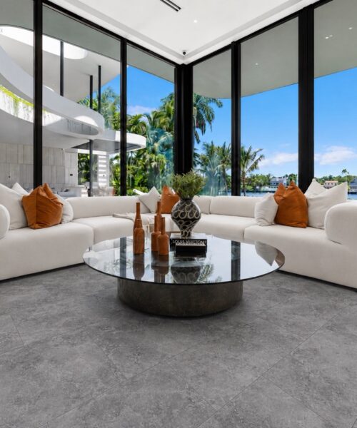 Midnight Slate stone-look flooring with subtle gray mineral variation surrounds curved white sofas in a bright contemporary living room with glass walls.