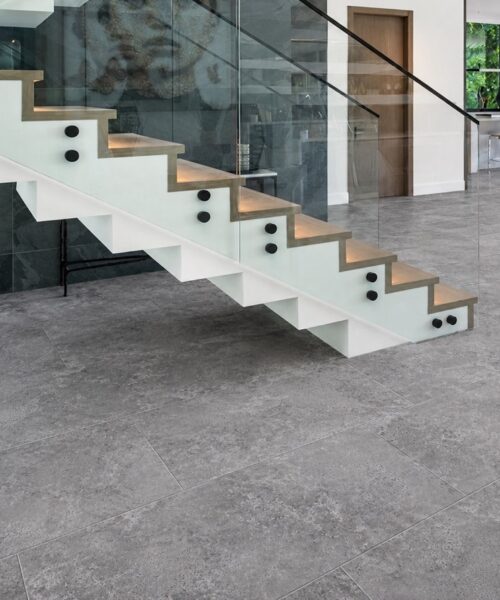 Midnight Slate slate-look flooring with visible mineral variation extends beneath a modern floating staircase with glass railing and warm wood treads.
