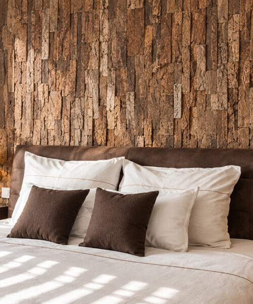 Stacked Stone - 55/64" (22mm) - Acoustic Cork Wall Tile (WSkSt22) Textured 3D cork wall panels in earthy brown tones create a warm bedroom feature wall behind a leather headboard.