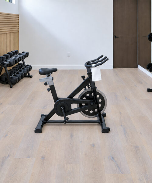 Nordic Oak 9.5mm waterproof Natural Composite Flooring shows pale plank variation beneath a stationary bike and weights.